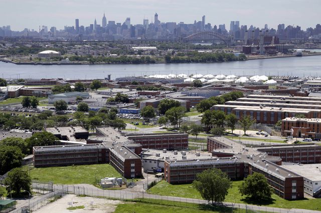 An aerial picture of Rikers Island jail facilities in the foreground, with the Manhattan skyline in the back